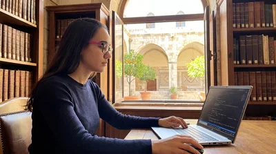 A young Syrian engineer at a desk in Damascus, notes and a laptop in front of her, late afternoon light through the window.
